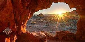 Dazzling ray of sun, seen through a red rock cavity, dances across a barren landscape at sunrise.